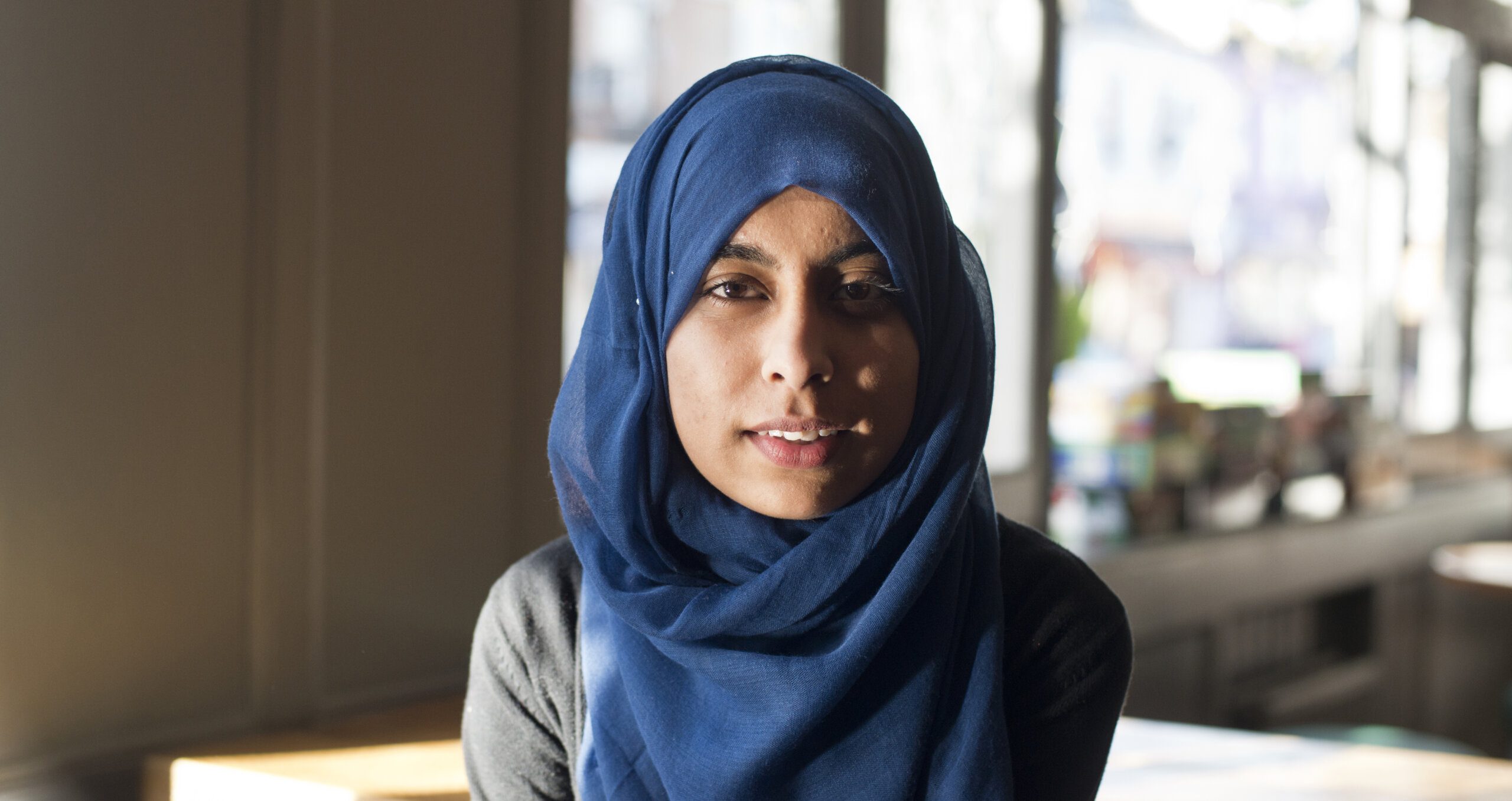 A woman wearing a blue hijab and a gray K-10 top sits indoors, looking at the camera with a soft expression. Sunlight streams through large windows in the background, creating a warm and inviting atmosphere.