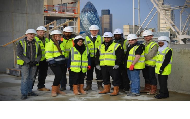 A group of K-10 construction workers in high-visibility jackets, hard hats, gloves, and boots stand together on a building site, with city skyscrapers visible in the background.