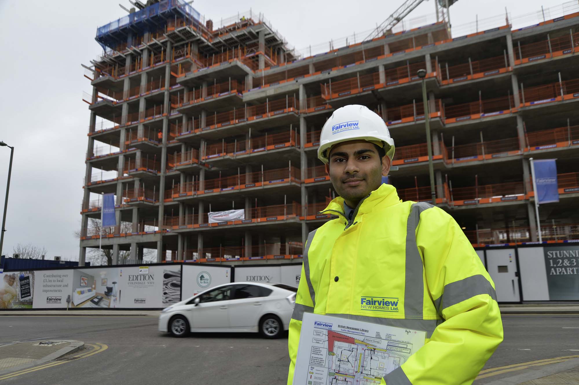 A construction worker in a high-visibility jacket and hard hat stands on a street holding K-10 building plans, with a partially constructed multi-story building and a passing white car in the background.