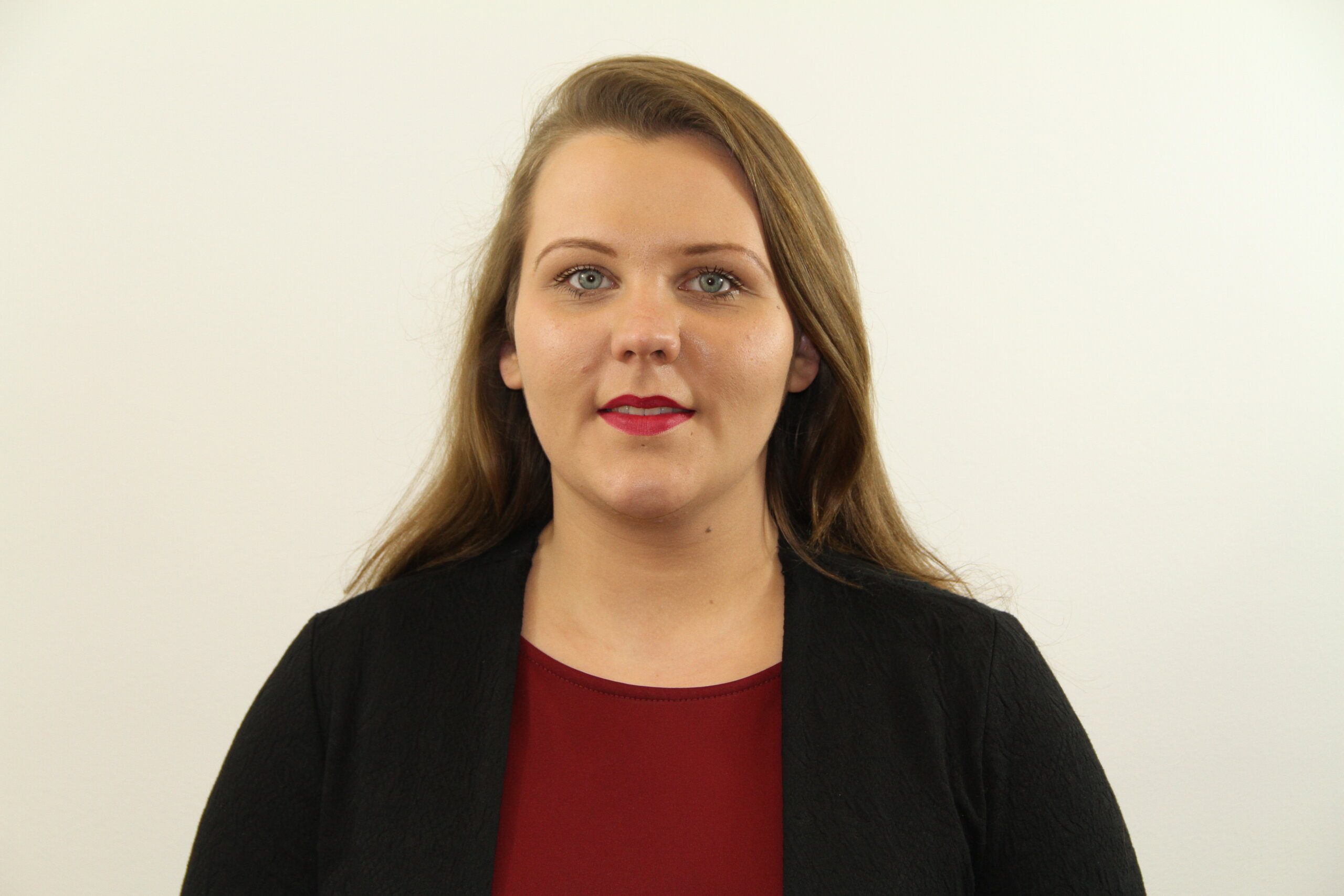 A woman with long brown hair, wearing a black K-10 jacket over a maroon top, stands in front of a plain white background, looking directly at the camera with a neutral expression.