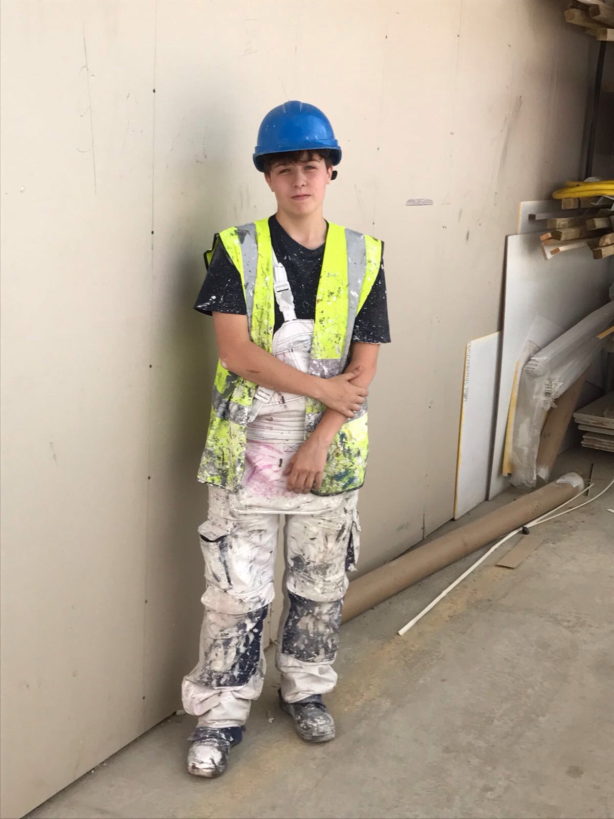 A young K-10 construction worker wearing a blue hard hat, yellow safety vest, and paint-splattered overalls stands against a wall in a workshop or construction site, surrounded by building materials.