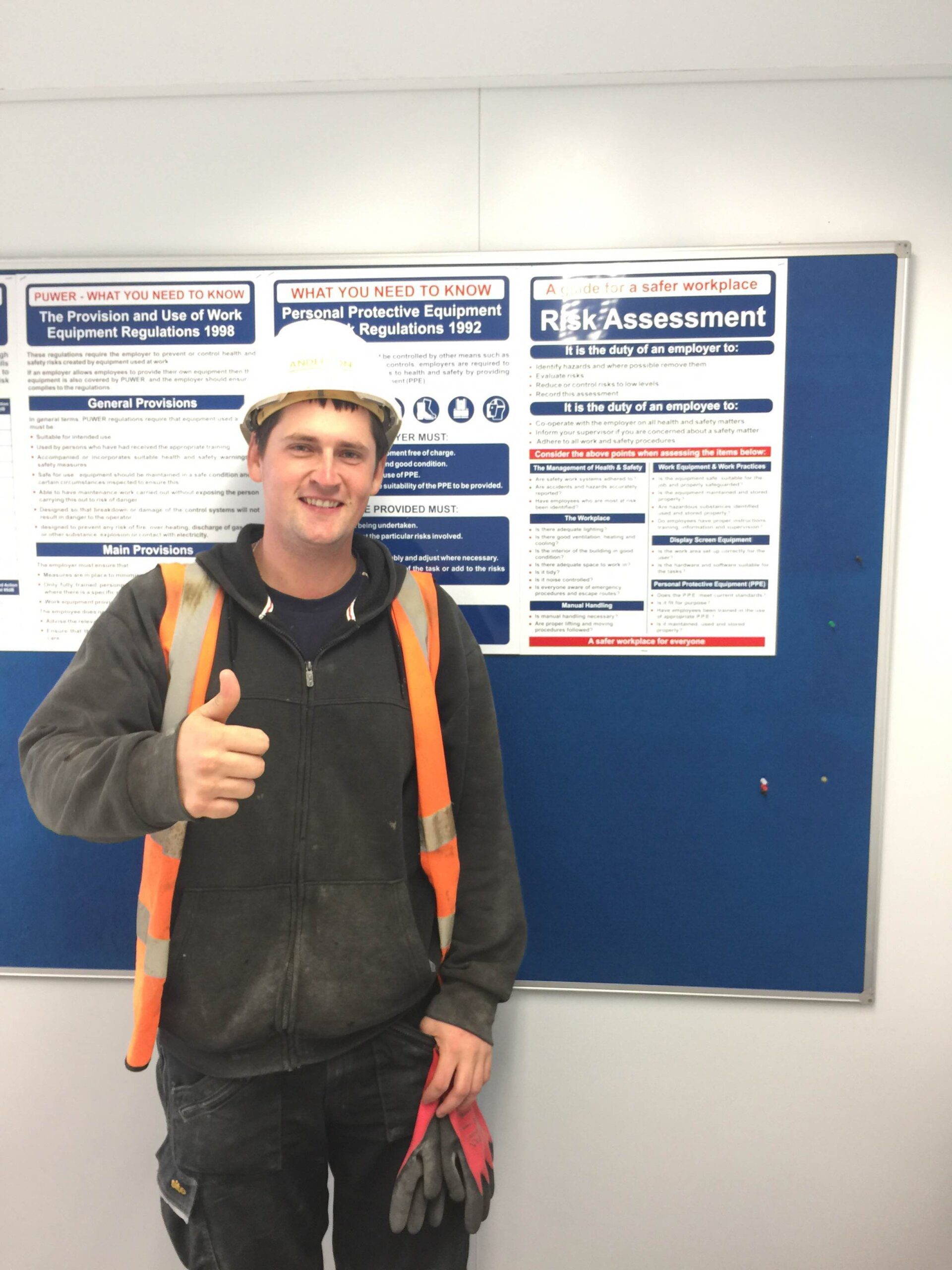 A construction worker in a hard hat and high-visibility vest stands indoors, smiling and giving a thumbs-up in front of a blue K-10 bulletin board displaying workplace safety and risk assessment posters.