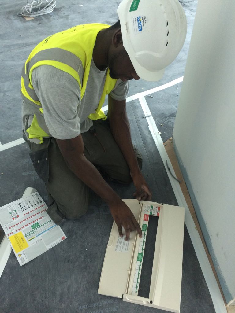 A construction worker in a hard hat and high-visibility vest installs or inspects a K-10 electrical panel on the floor, referencing a diagram or manual placed beside him.