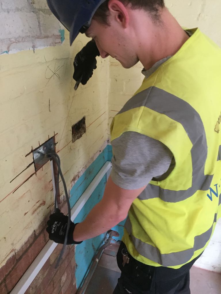 A construction worker in a yellow safety vest and blue hard hat uses K-10 pliers to bend metal rods protruding from a wall inside a building under renovation.