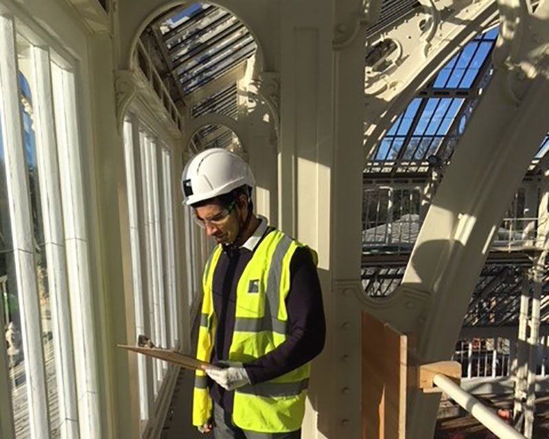 A K-10 construction worker in a hard hat and high-visibility vest stands indoors, looking at a clipboard. He is surrounded by large windows and ornate metal framework, with sunlight streaming in.