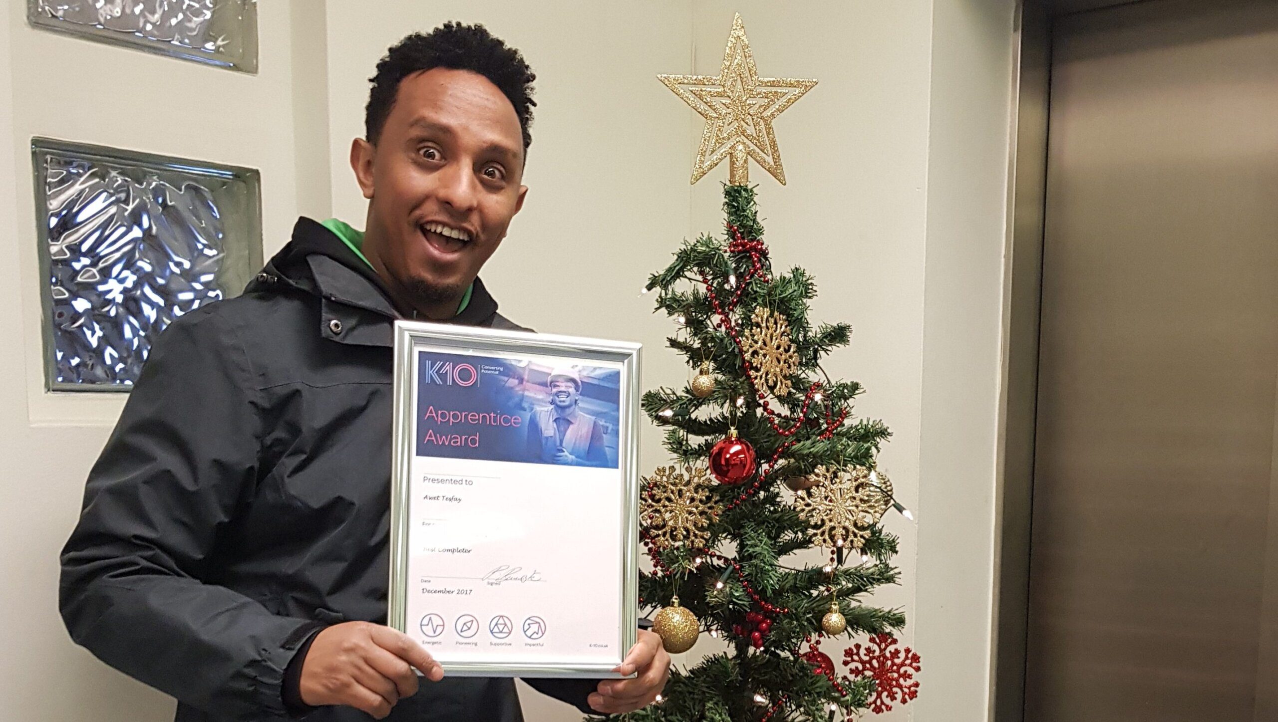A smiling man wearing a dark jacket holds a K-10 apprentice award certificate while standing next to a decorated Christmas tree with a gold star topper.