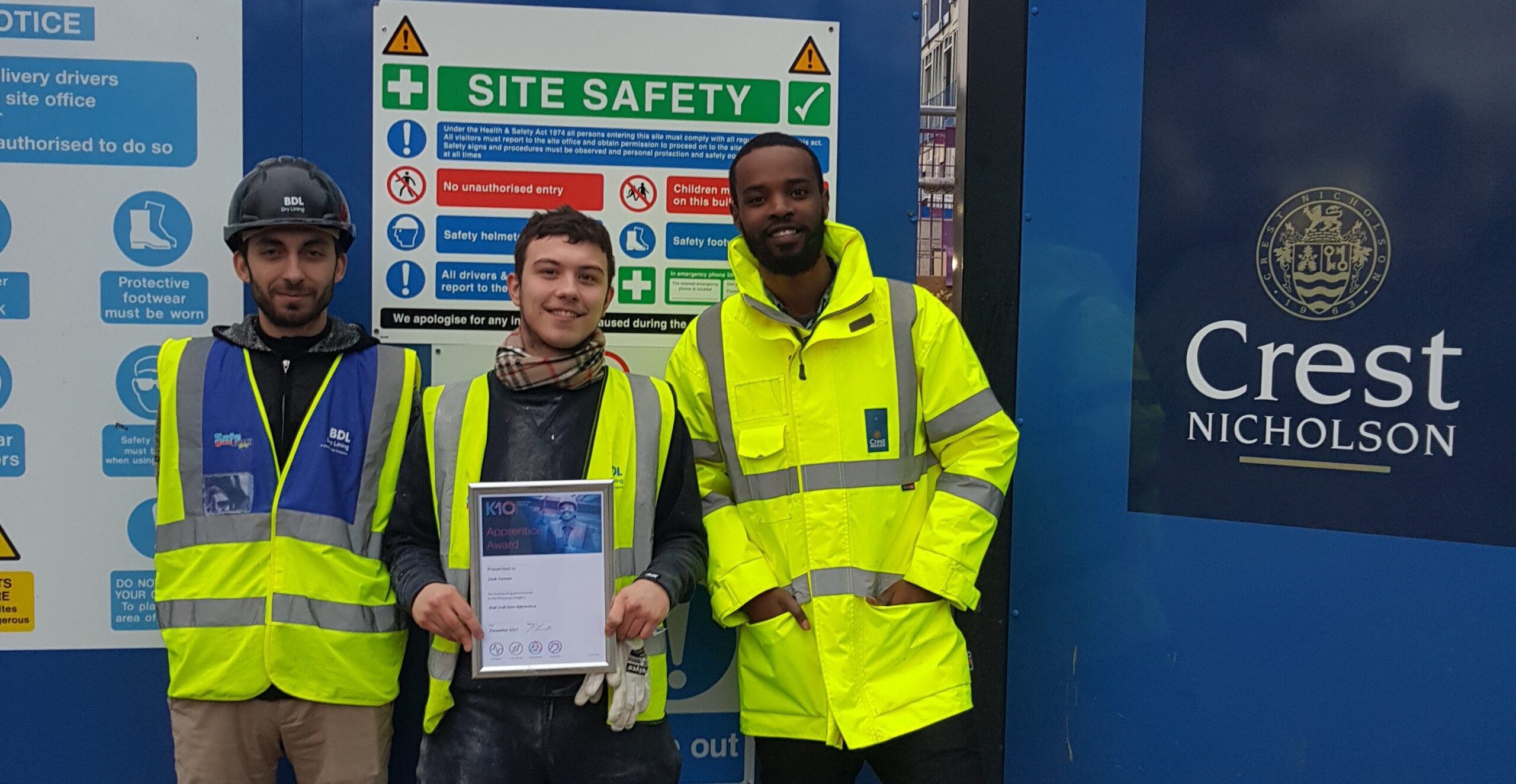 Three K-10 construction workers in safety gear pose and smile together at a construction site. One proudly holds a certificate, while safety signs and a Crest Nicholson logo are displayed on the wall behind them.