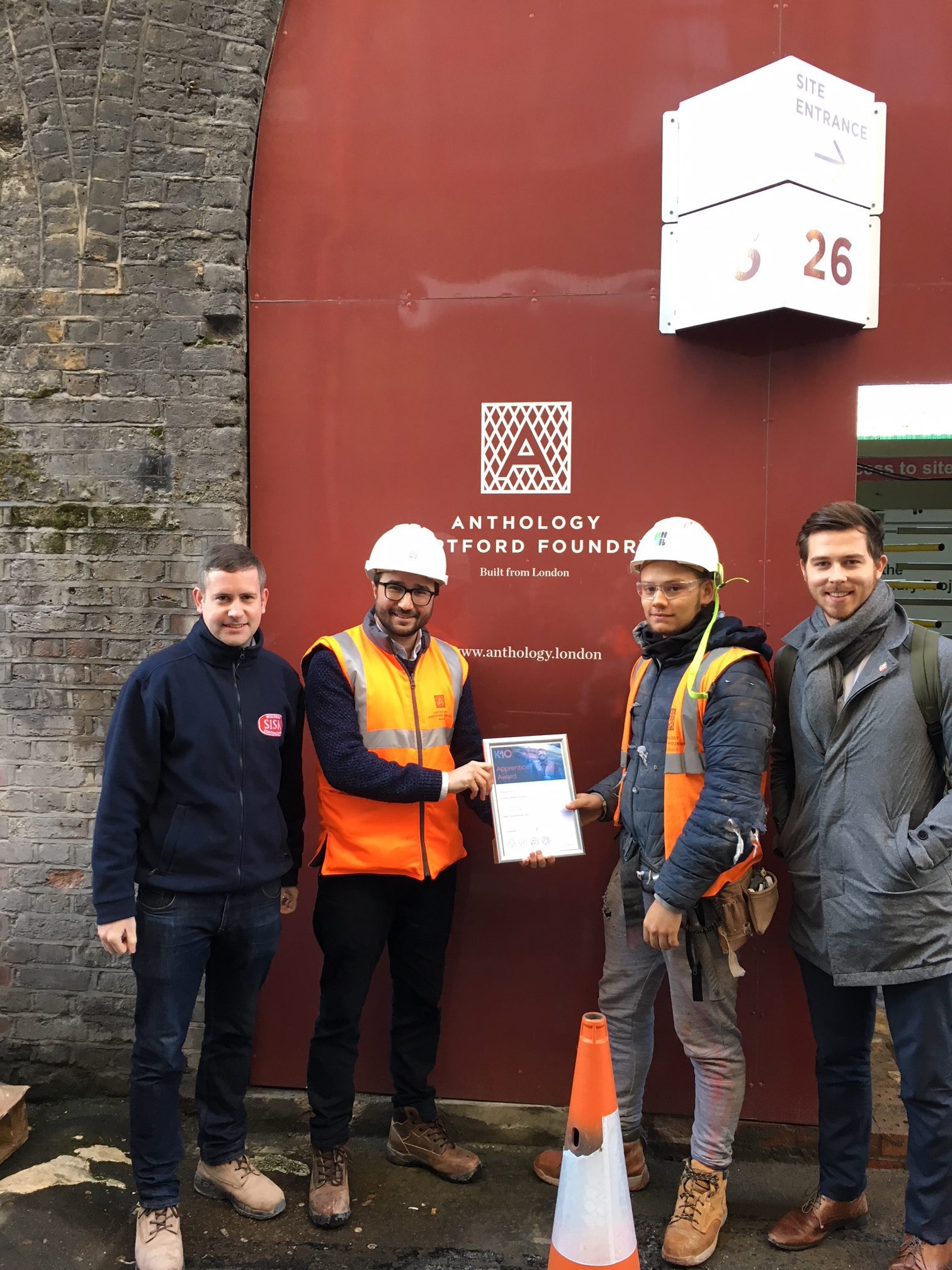 Four men stand in front of a red wall with the Anthology Deptford Foundry logo. Two wear K-10 construction gear and hold a certificate; the others are in casual outfits. A traffic cone is in front.