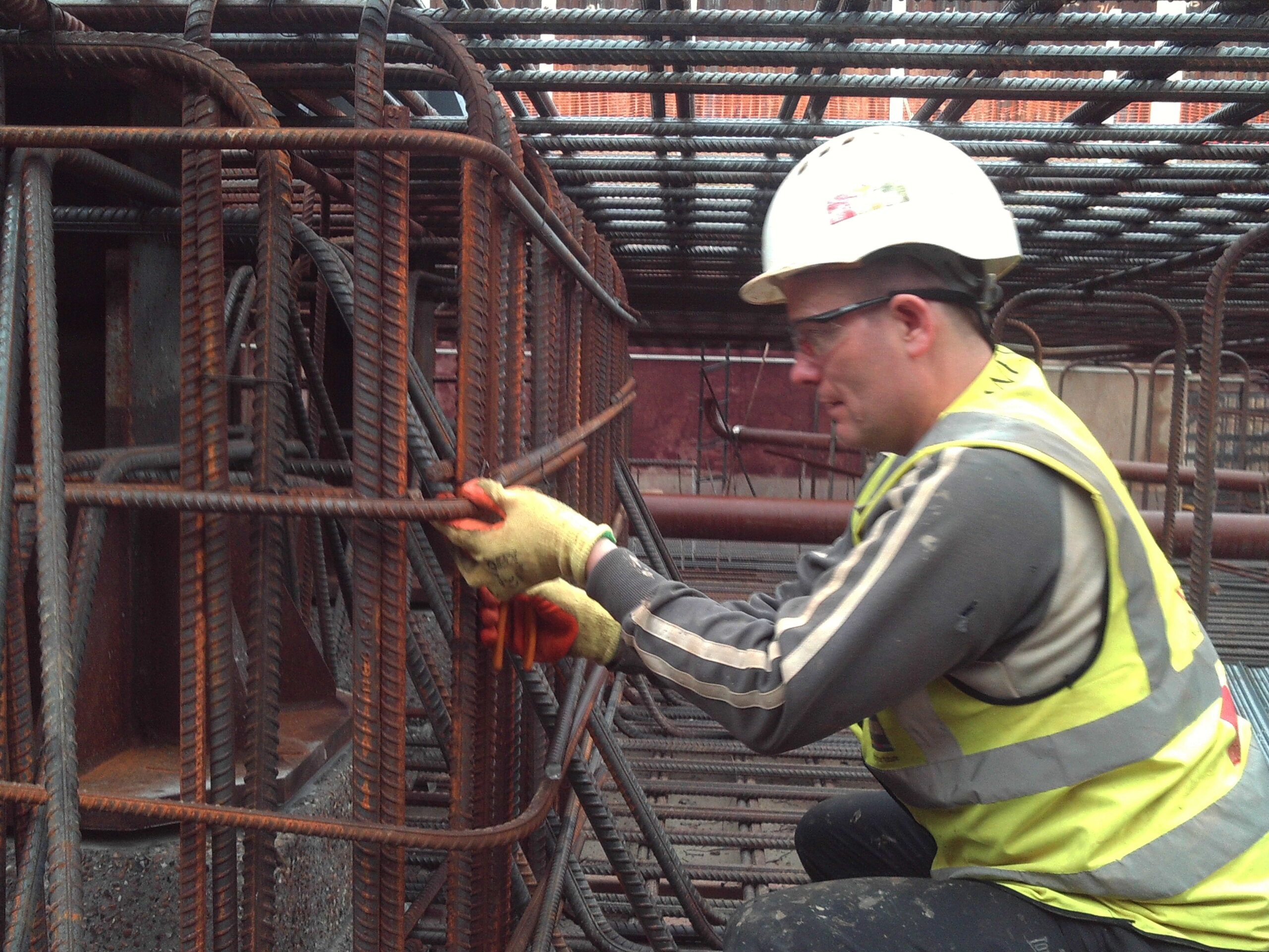A construction worker wearing a K-10 hard hat, safety glasses, gloves, and a high-visibility vest is tying steel rebar together on a construction site.