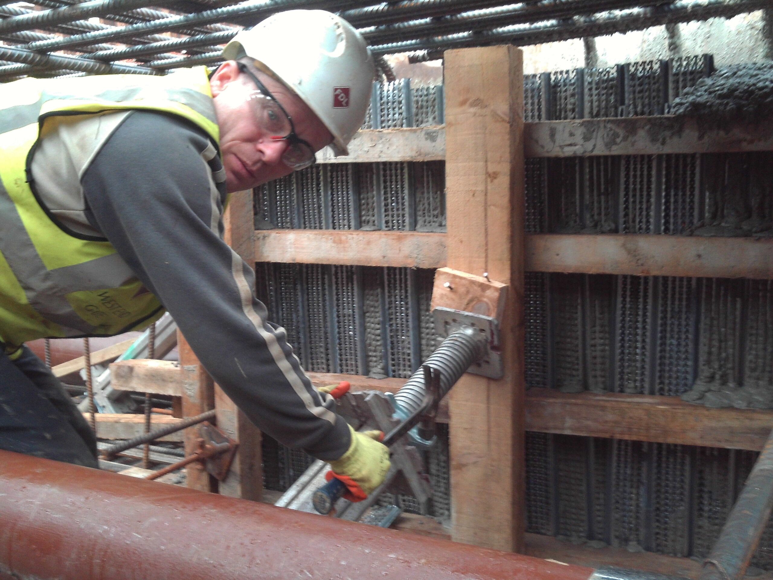 A construction worker wearing a hard hat, safety glasses, and a high-visibility vest uses a large K-10 wrench to tighten a metal rod against wooden and steel reinforced formwork at a construction site.