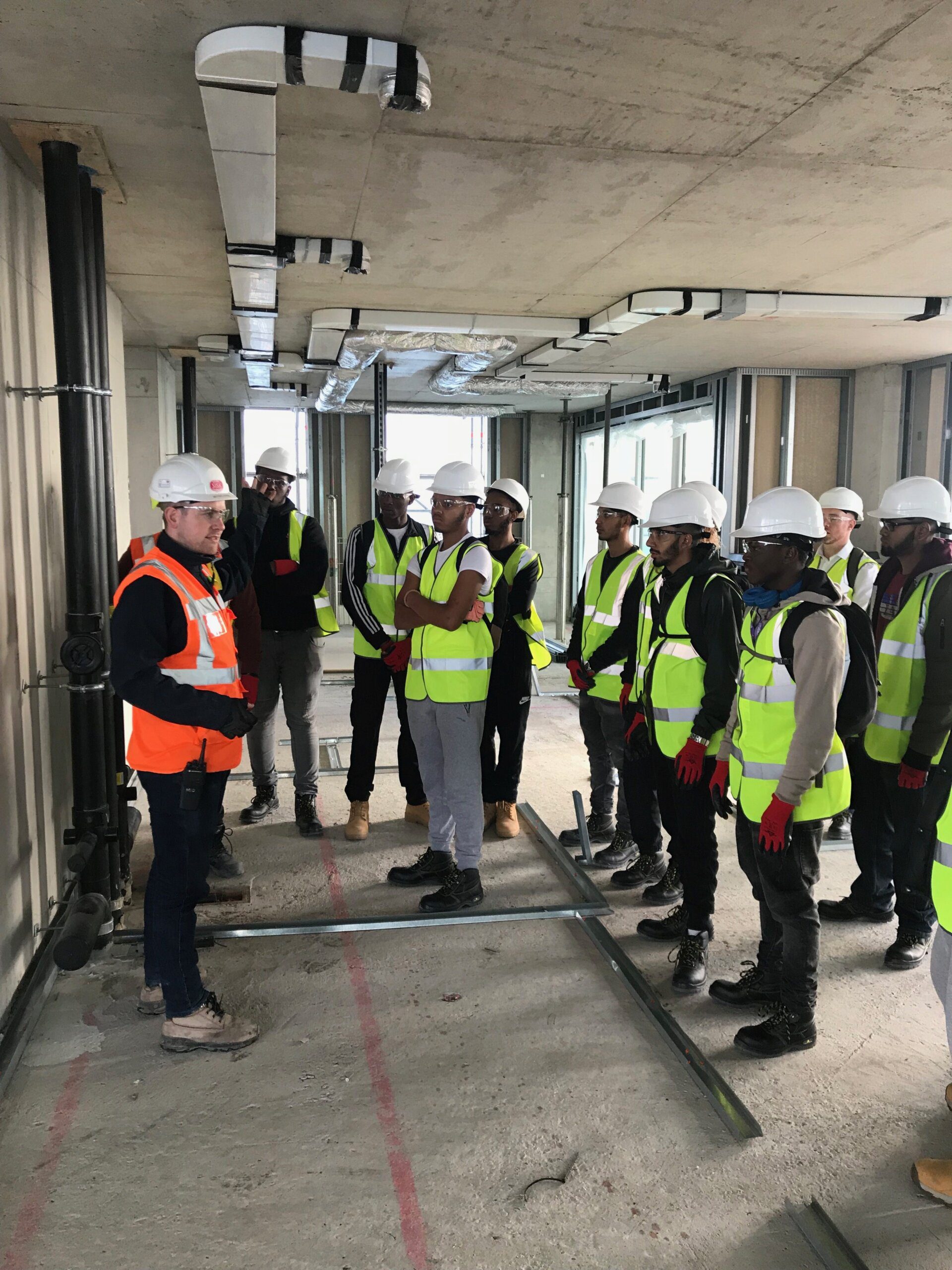 A group of people wearing hard hats, reflective vests, and gloves listen to a construction worker giving K-10 safety instructions inside a building under construction. Exposed pipes and concrete surfaces are visible.