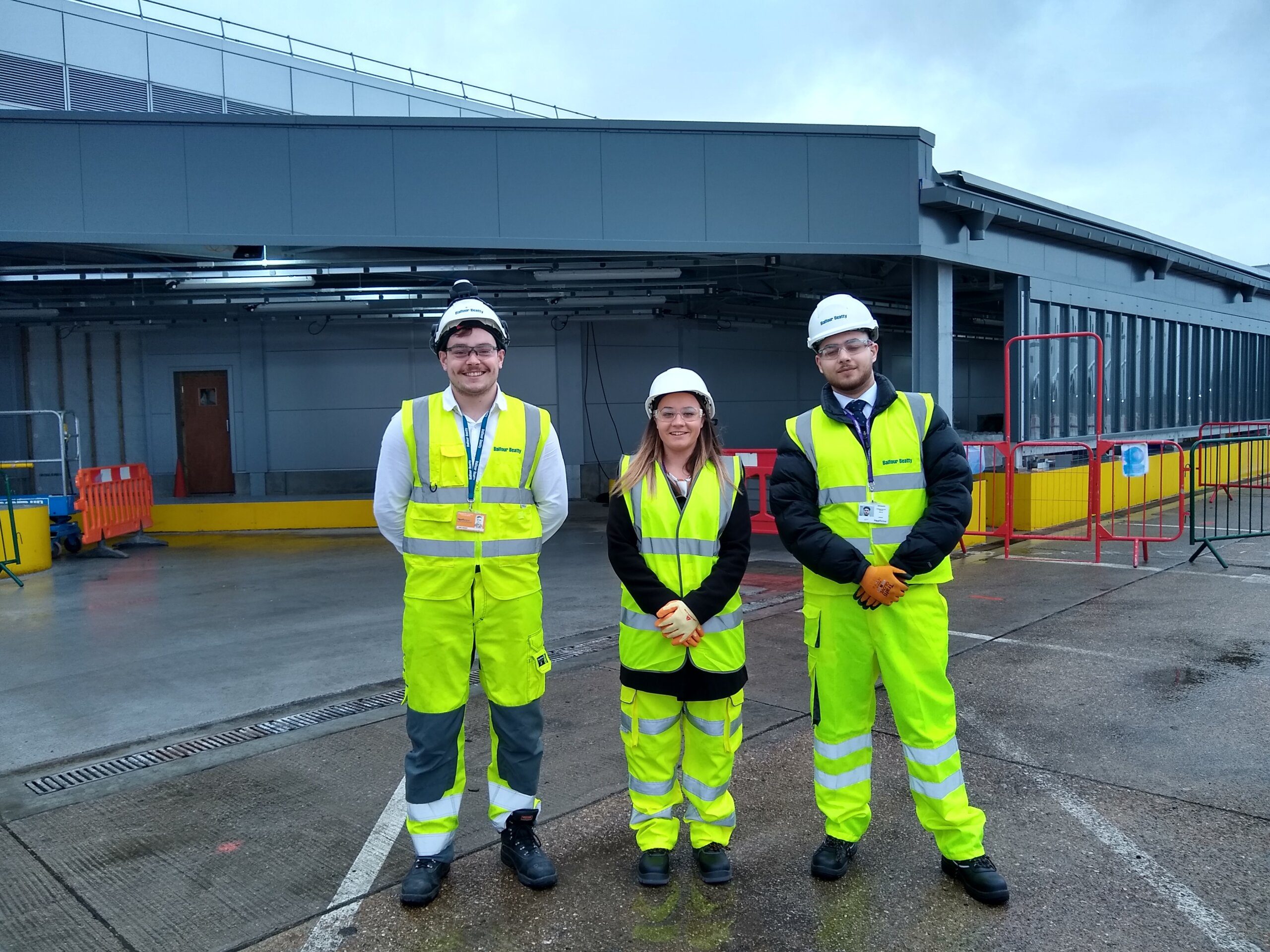 Three people in yellow high-visibility jackets, hard hats, and safety gear stand outdoors beside a K-10 building under construction, with barriers and construction materials visible nearby.