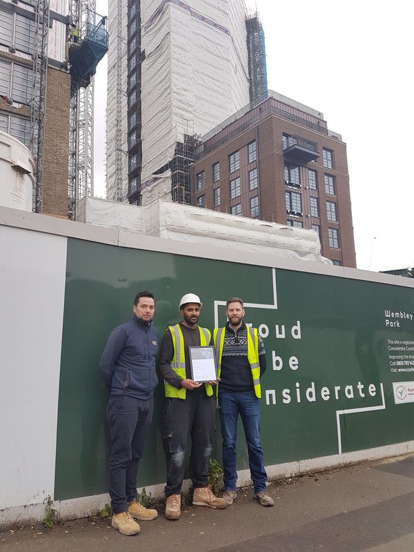 Three men, including one in construction gear holding a K-10 certificate, stand in front of a green construction site wall with partially visible text. A tall building under construction rises in the background.