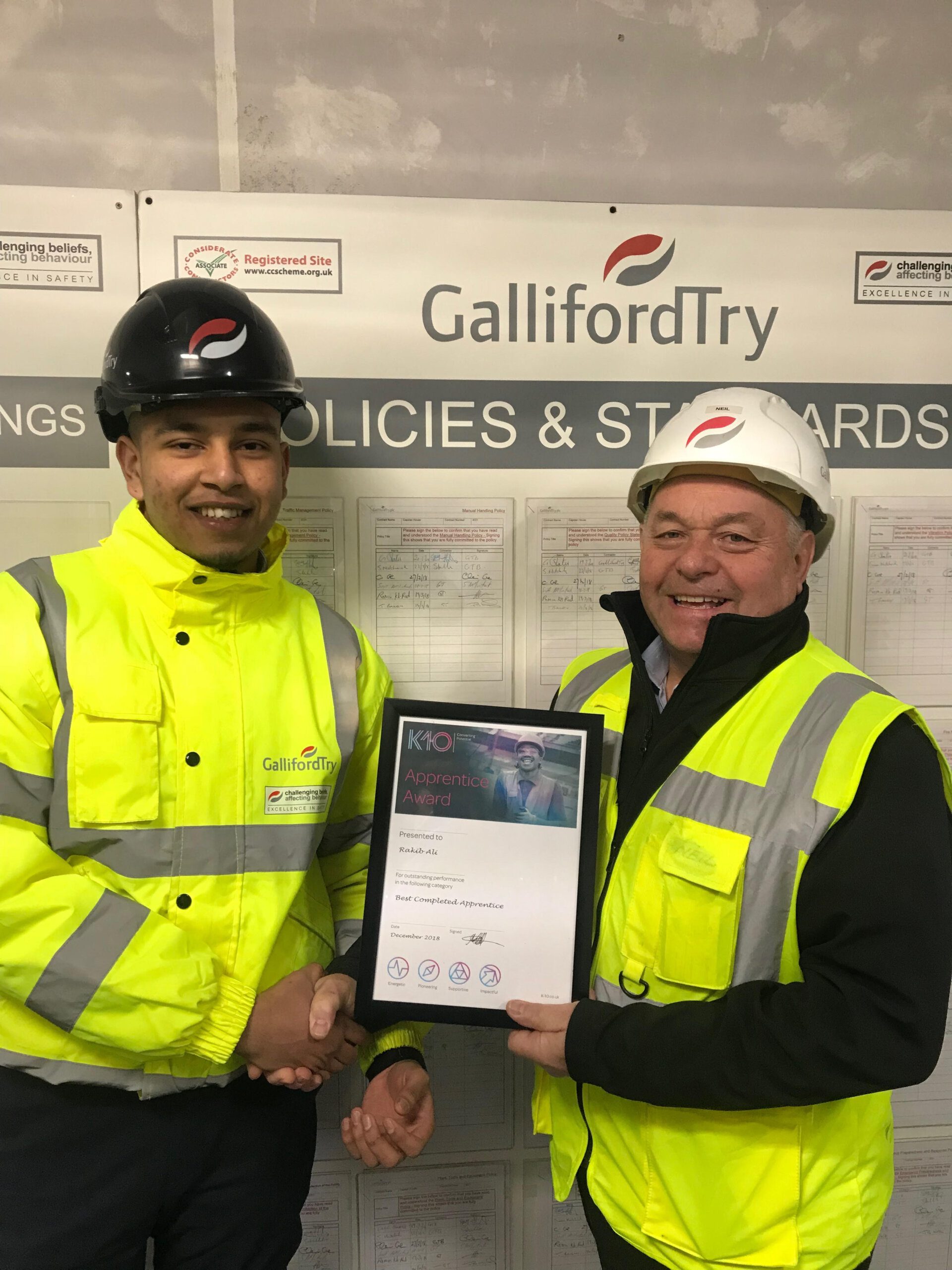 Two men in high-visibility jackets and hard hats smile while shaking hands—one holds a K-10 Apprentice Award certificate. They stand in front of a Galliford Try banner and site notices.