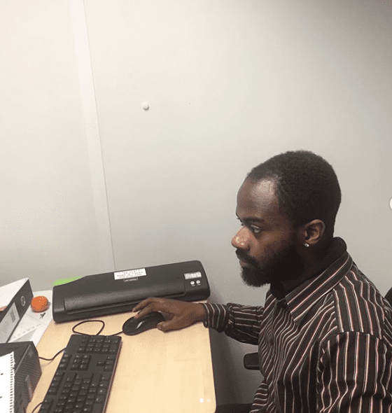 A man with a beard sits at a desk using a computer mouse and keyboard, focused on his K-10 work. He is wearing a striped shirt and looking at a monitor. Office supplies, including a laminator and an orange, are on the desk.