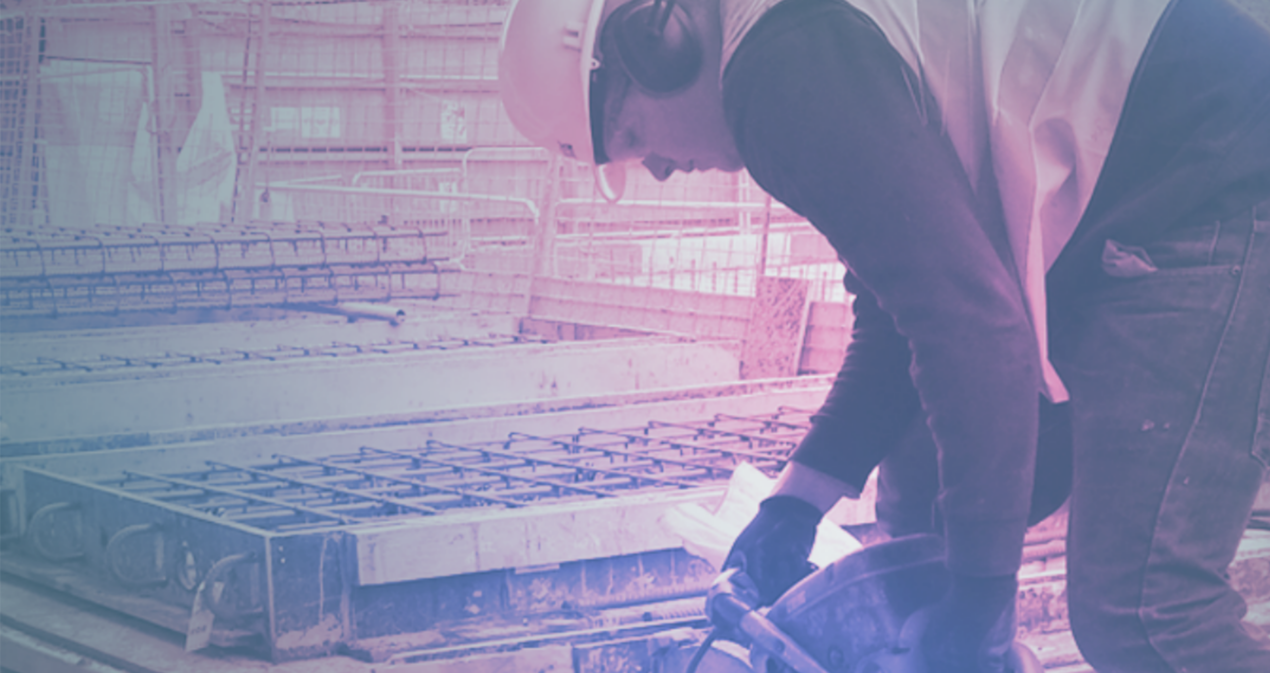 A construction worker in a hard hat and safety gear bends over to use a K-10 power tool at a building site, surrounded by metal rebar and concrete structures.
