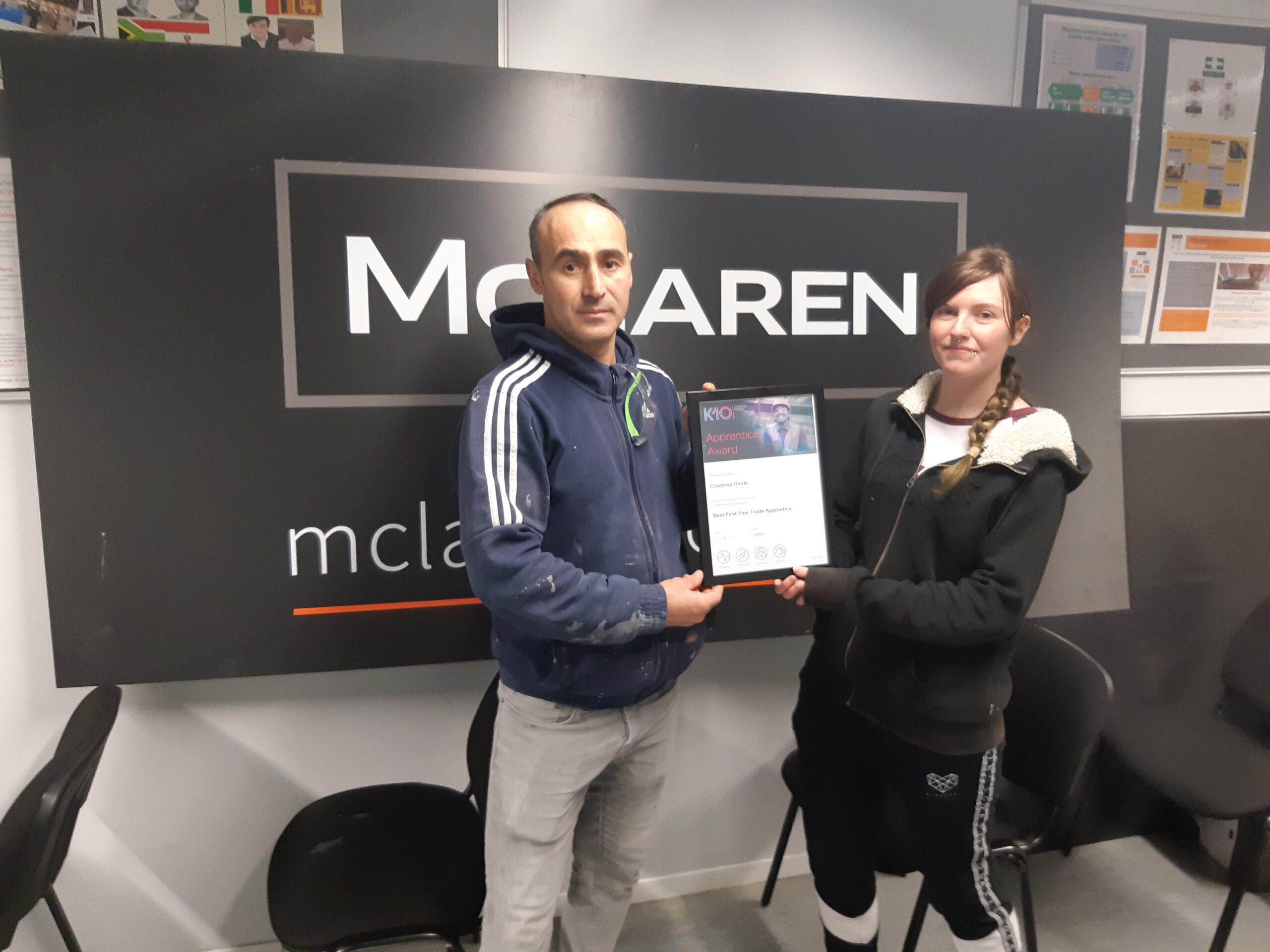 Two people stand indoors in front of a McLaren sign, holding a K-10 certificate together and facing the camera. They both appear to be smiling slightly and are wearing casual clothing.