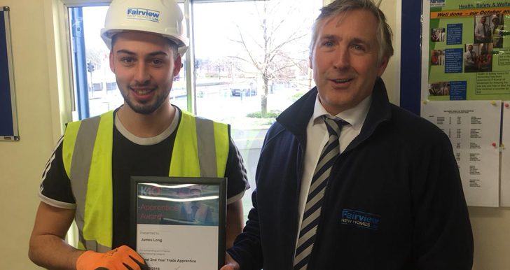 Two men stand indoors. The younger man, in a high-visibility vest and gloves, holds a K-10 Apprentice Award certificate. The older man beside him smiles, both appearing pleased. Company branding says Fairview New Homes.