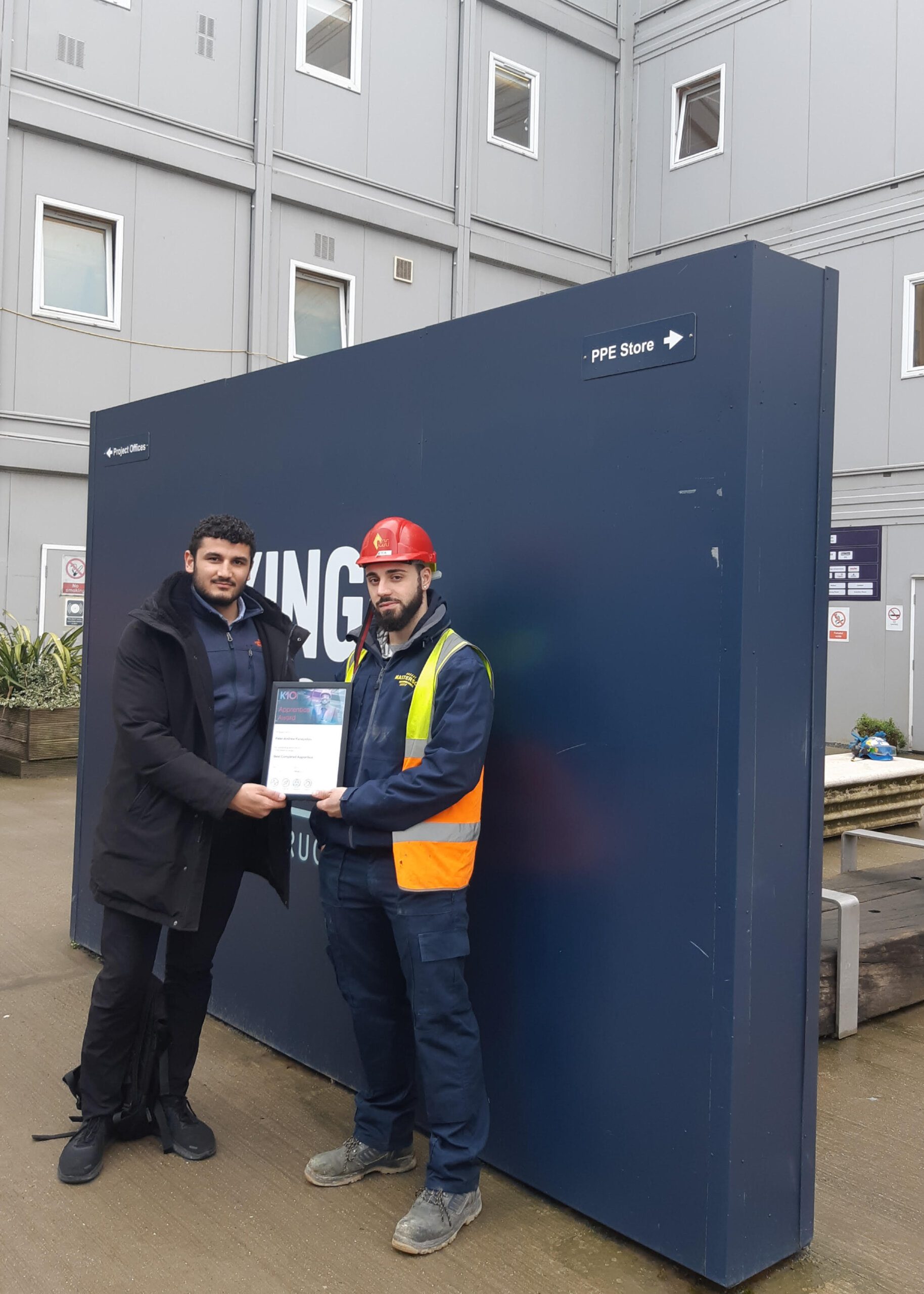 Two men stand in front of a dark blue wall outside an industrial building; one is wearing a black coat and the other, in K-10 safety gear—a hard hat, vest, and work clothes. They hold a certificate together and face the camera.