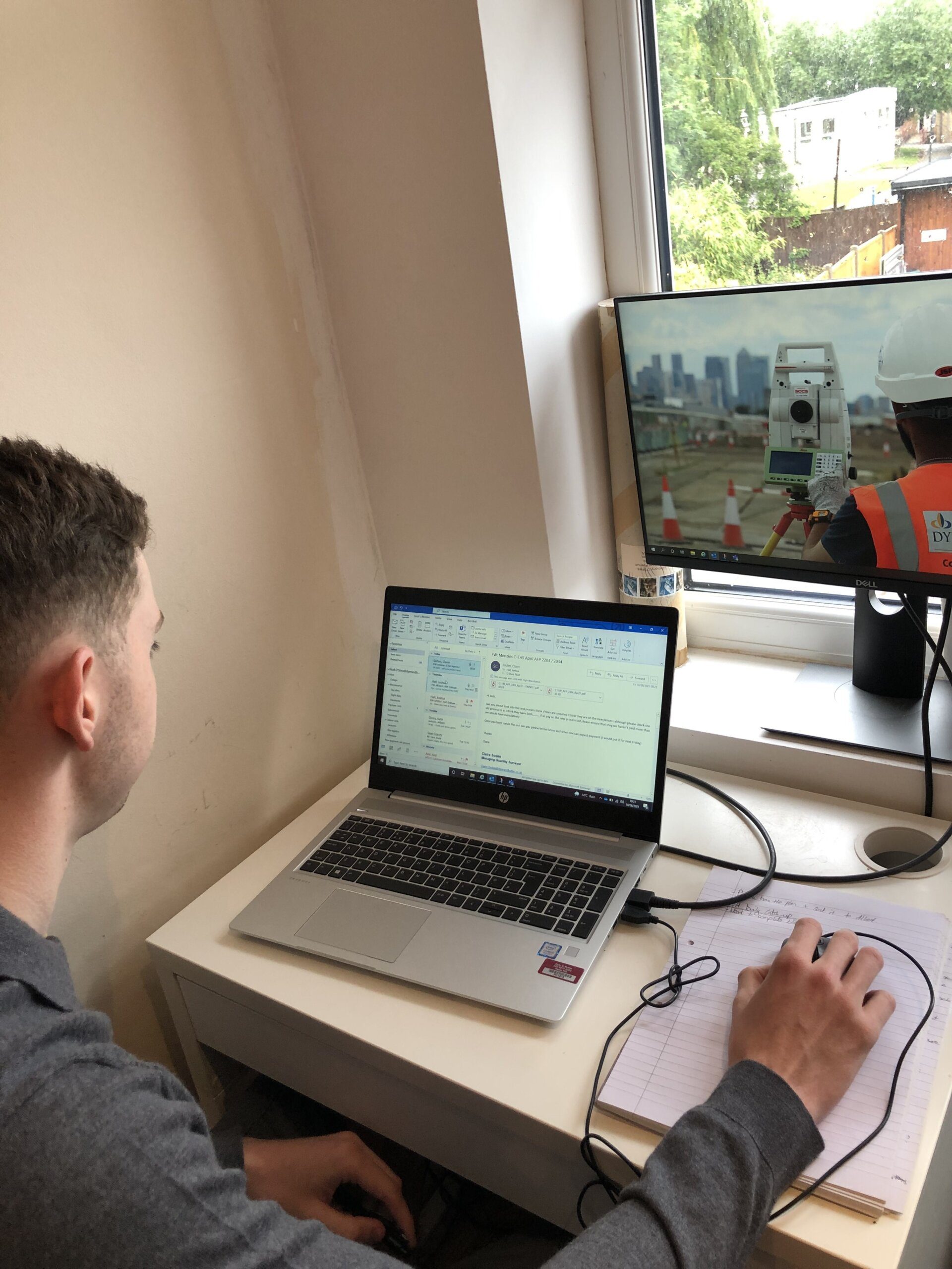 A person sits at a desk using a laptop with an email inbox open. A monitor nearby displays a K-10 safety vest-clad worker using surveying equipment outdoors. A notepad and pen are also on the desk.