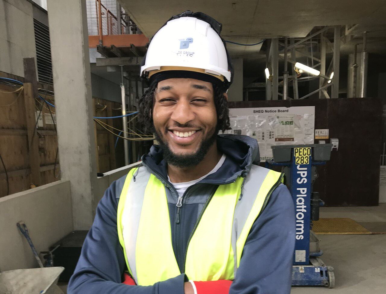 A smiling construction worker wearing a white hard hat, neon yellow K-10 safety vest, and dark hoodie stands with arms crossed at a construction site with equipment and signage in the background.