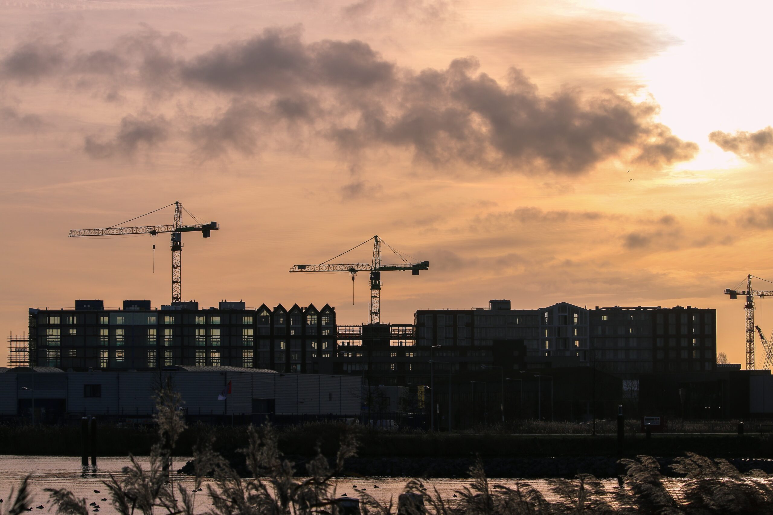 Silhouettes of construction cranes and buildings, including a K-10 model, stand against a cloudy, golden sunset sky. Grassy plants and water in the foreground hint at a developing urban area.