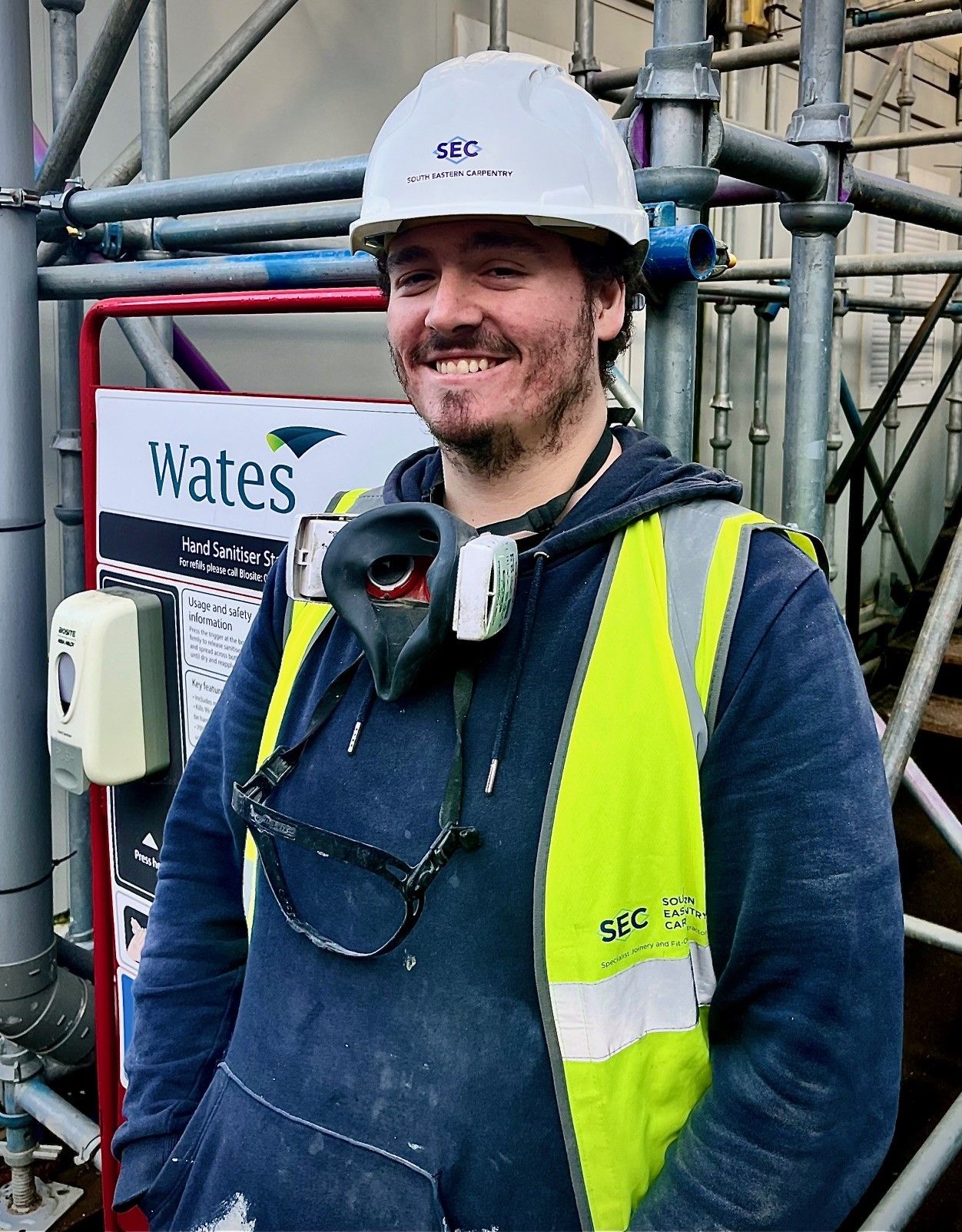 A construction worker wearing a white helmet, yellow safety vest, and blue hoodie stands in front of K-10 scaffolding. He is smiling, with a respirator and safety glasses around his neck. A hand sanitizer station is behind him.