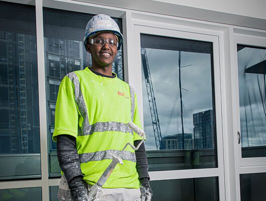 A construction worker in a K-10 hard hat, safety glasses, and a bright yellow high-visibility shirt stands in front of large glass windows with buildings and a crane in the background.