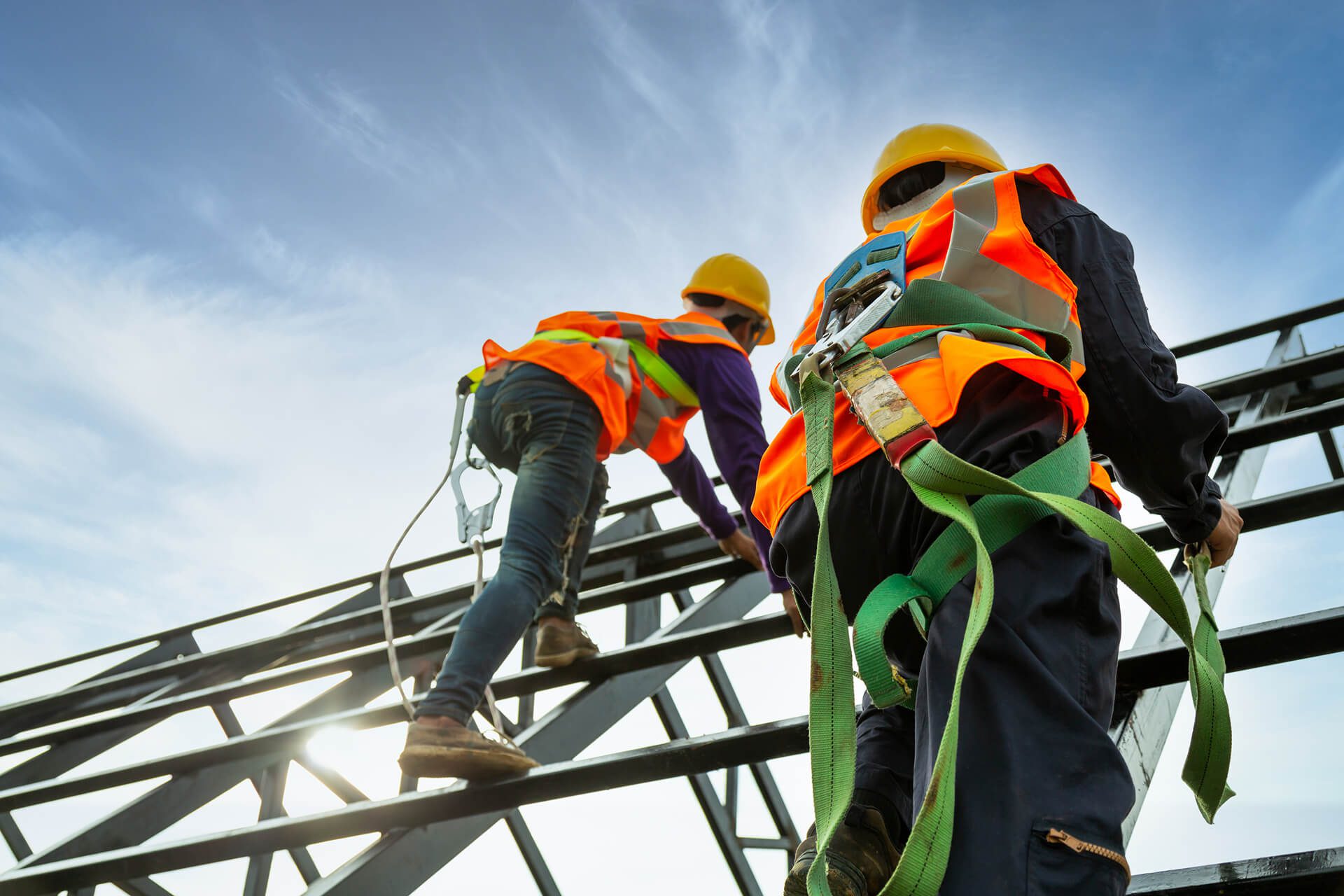 Two K-10 construction workers wearing safety harnesses, orange reflective vests, and yellow helmets climb a metal structure under a blue sky.